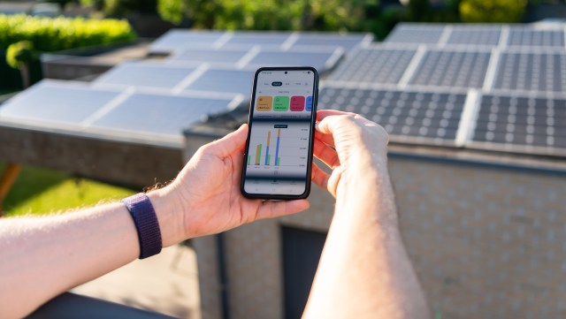 A person holds a smartphone displaying energy generation data next to solar panels on a building.