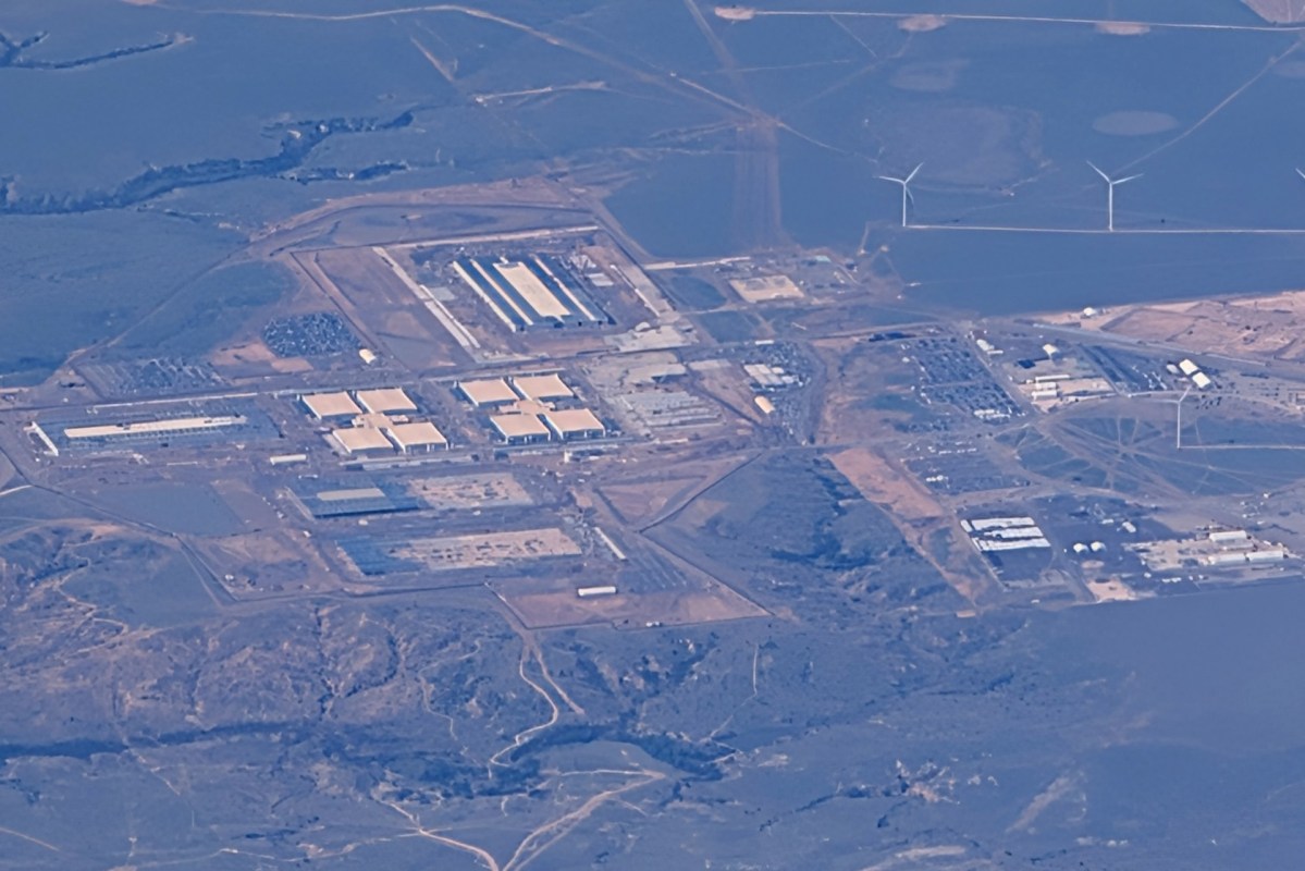 An aerial view of an industrial area with large buildings, highways, and wind turbines in the surrounding landscape.