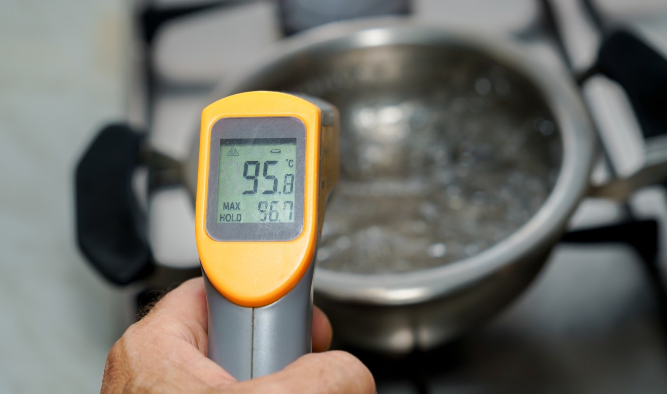 A thermometer displays a reading of 95.8°C next to a boiling pot of water on a stove.