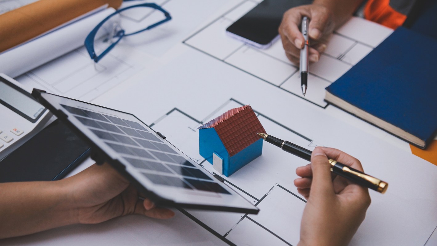 Hands holding a tablet and a pen, pointing at a small model house and a mock solar panel on architectural plans.