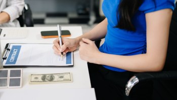 A person signs a contract on a clipboard with a solar panel, a dollar bill and a calculator on the table.