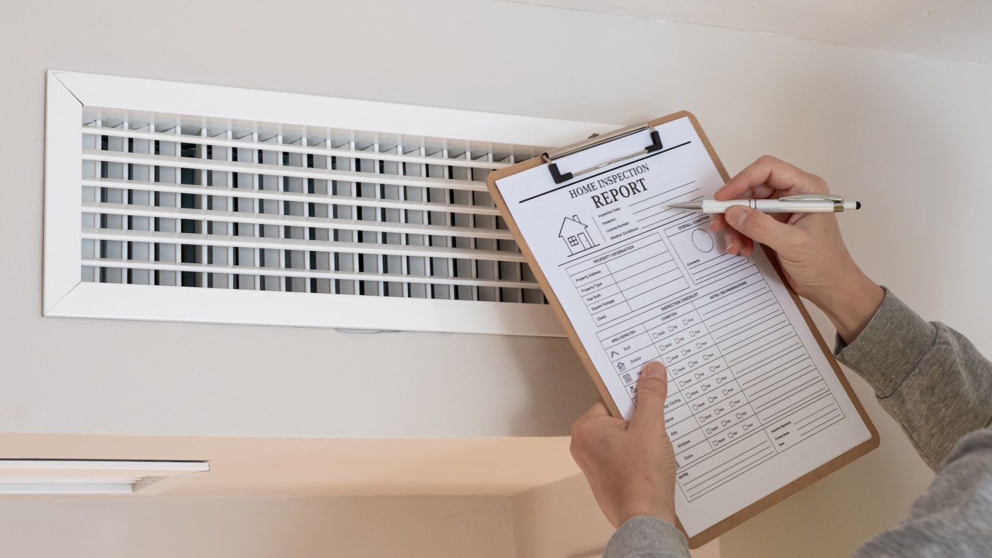 A person holds a clipboard with a home inspection report near a vent in a room.