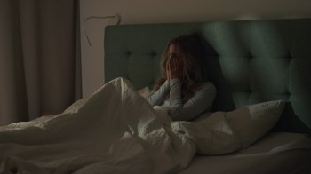 A woman sits on a bed, covering her face with her hands, surrounded by blankets and pillows.