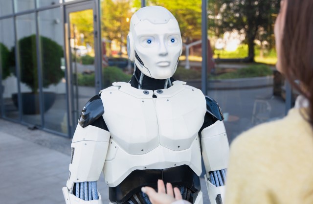 A robotic figure with a humanoid design interacts with a person outdoors near large glass windows.