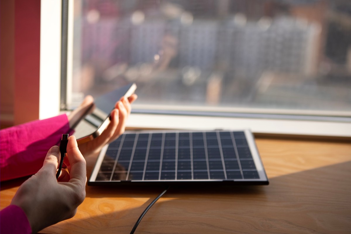 A person connects a smartphone to a solar panel while sitting by a window.