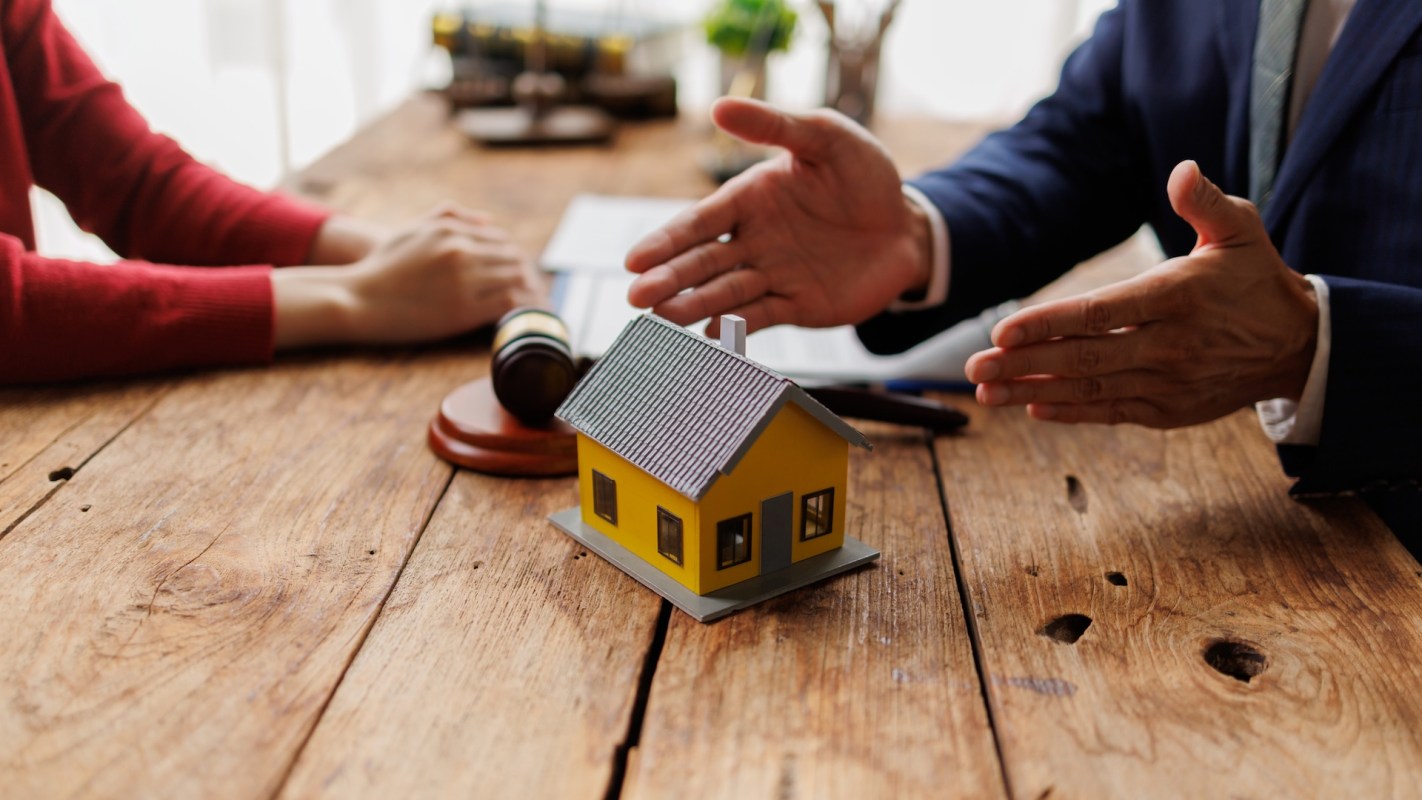 A miniature yellow house on a table with a gavel and two people discussing documents.