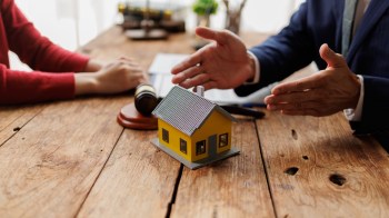A miniature yellow house on a table with a gavel and two people discussing documents.