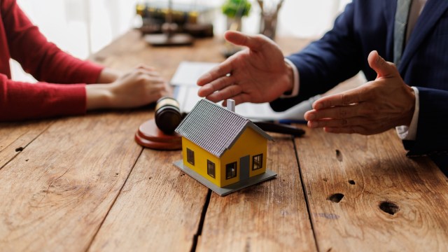 A miniature yellow house on a table with a gavel and two people discussing documents.