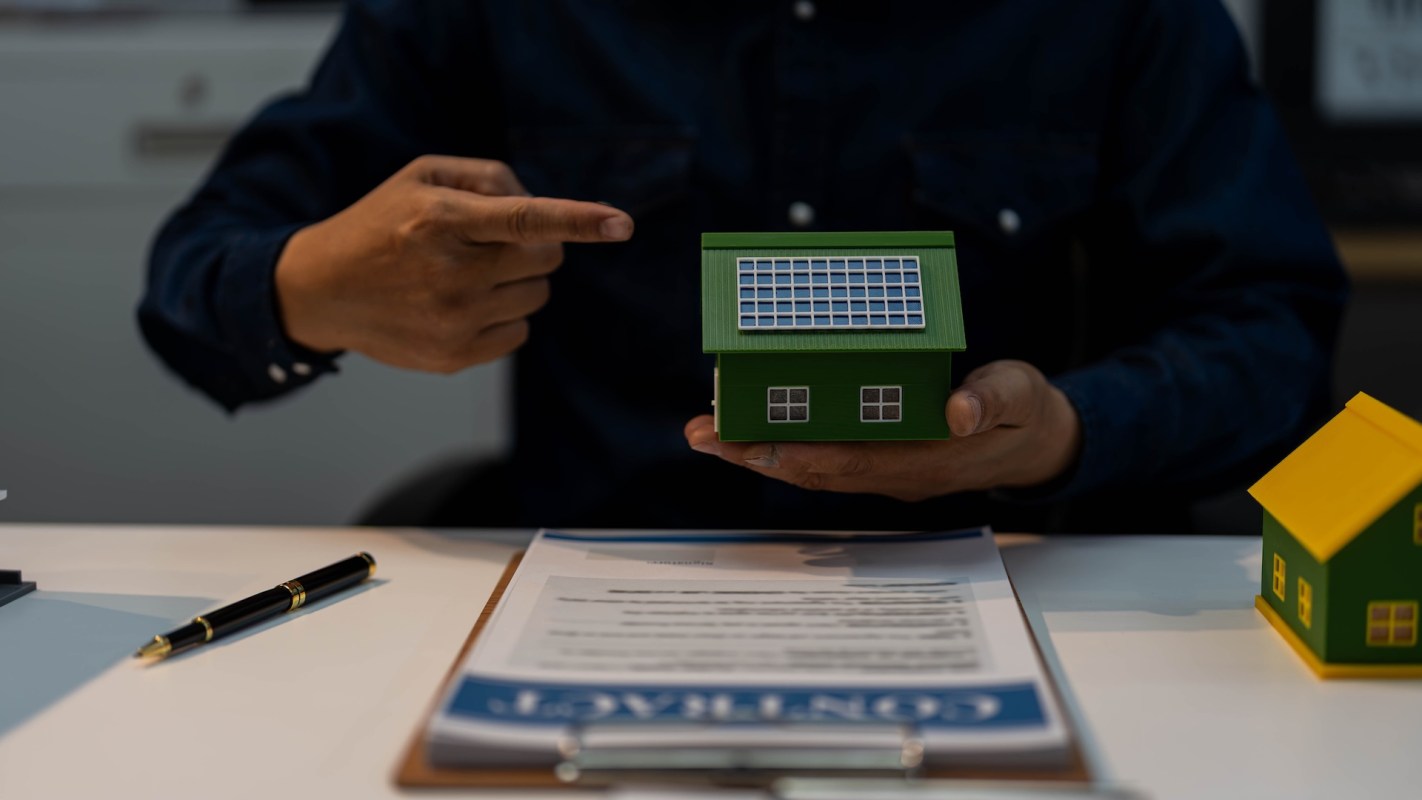 A person points to a model house with solar panels while sitting at a table with a contract.