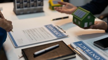 A person holds a model house with solar panels while discussing a contract on a table with papers and a pen.