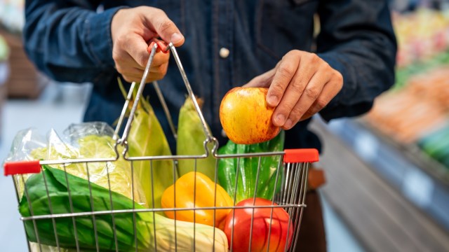A person places an apple into a shopping basket filled with various vegetables.