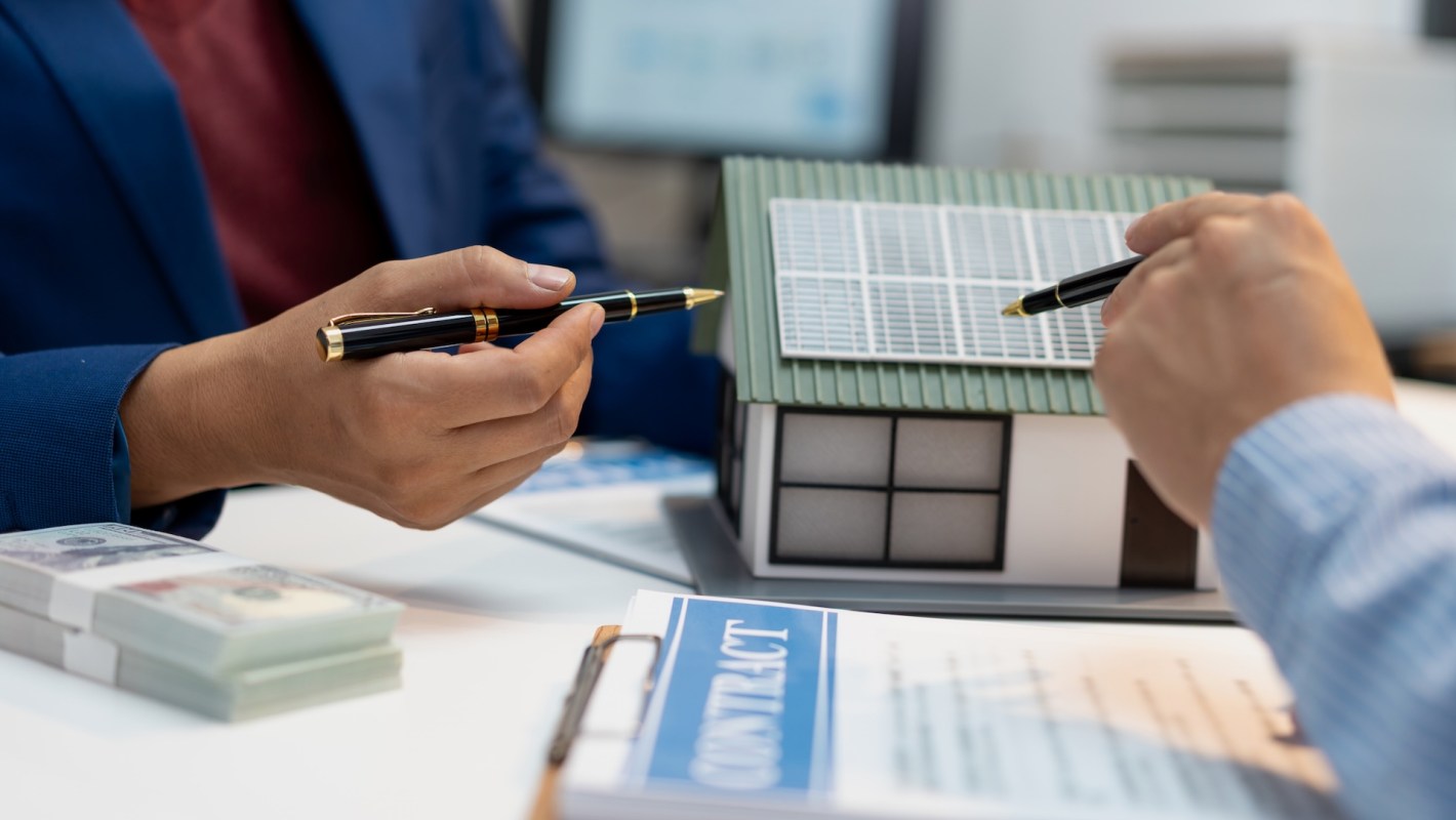 Two people discuss a house model with solar panels while reviewing a contract and stacks of money.