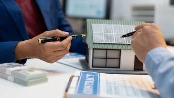 Two people discuss a house model with solar panels while reviewing a contract and stacks of money.