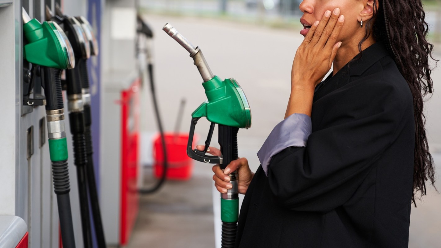 A woman holds a gas pump nozzle and touches her face while standing at a fuel station.