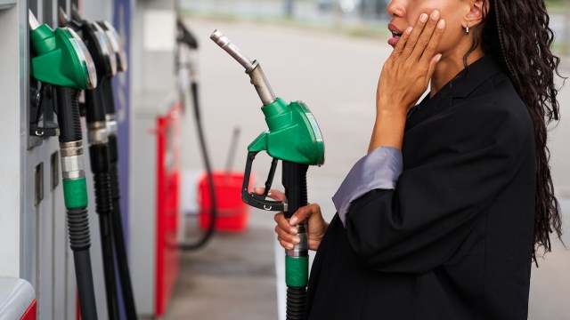 A woman holds a gas pump nozzle and touches her face while standing at a fuel station.