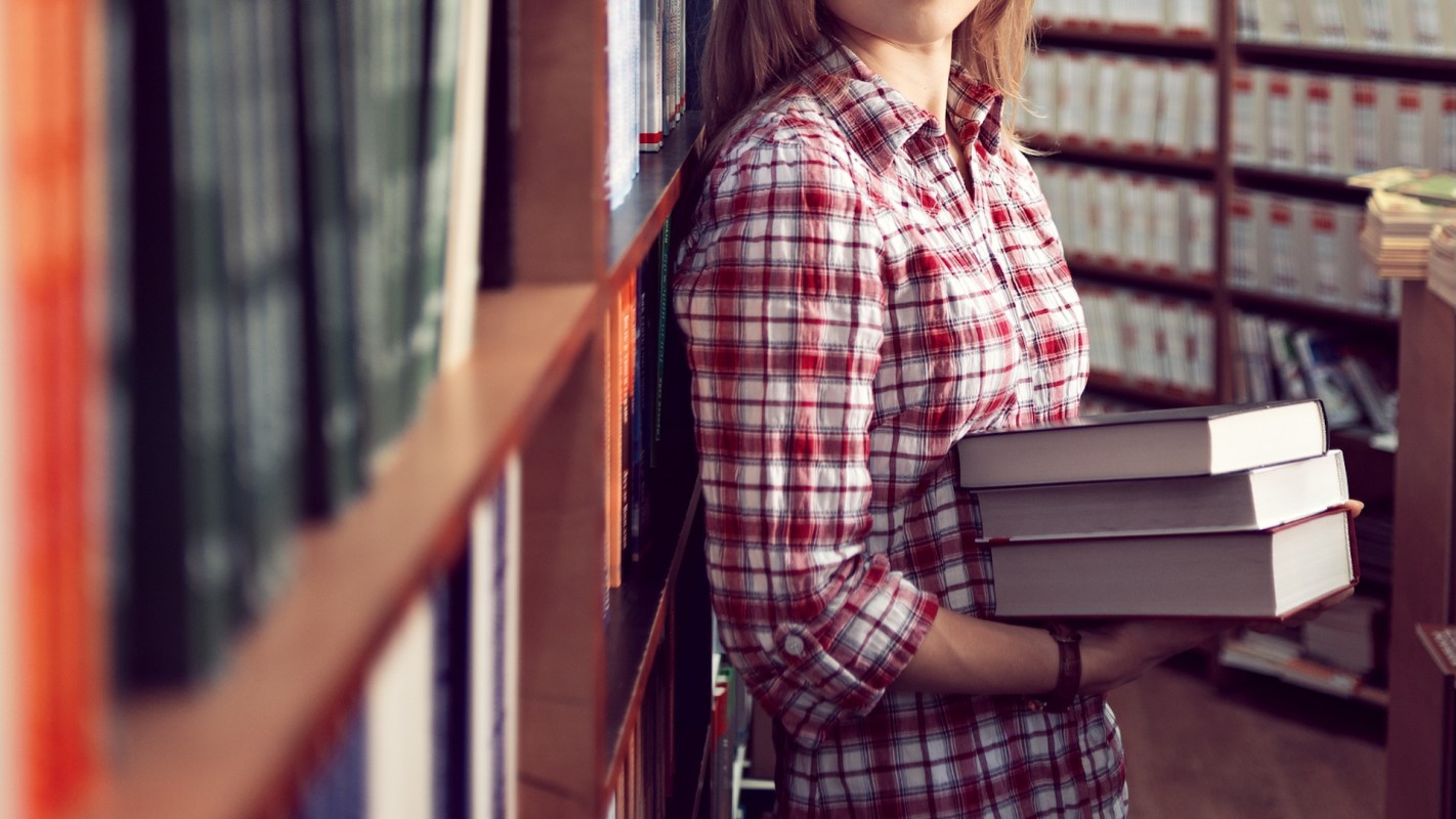 A book shop owner holds several books while standing in a stock room.