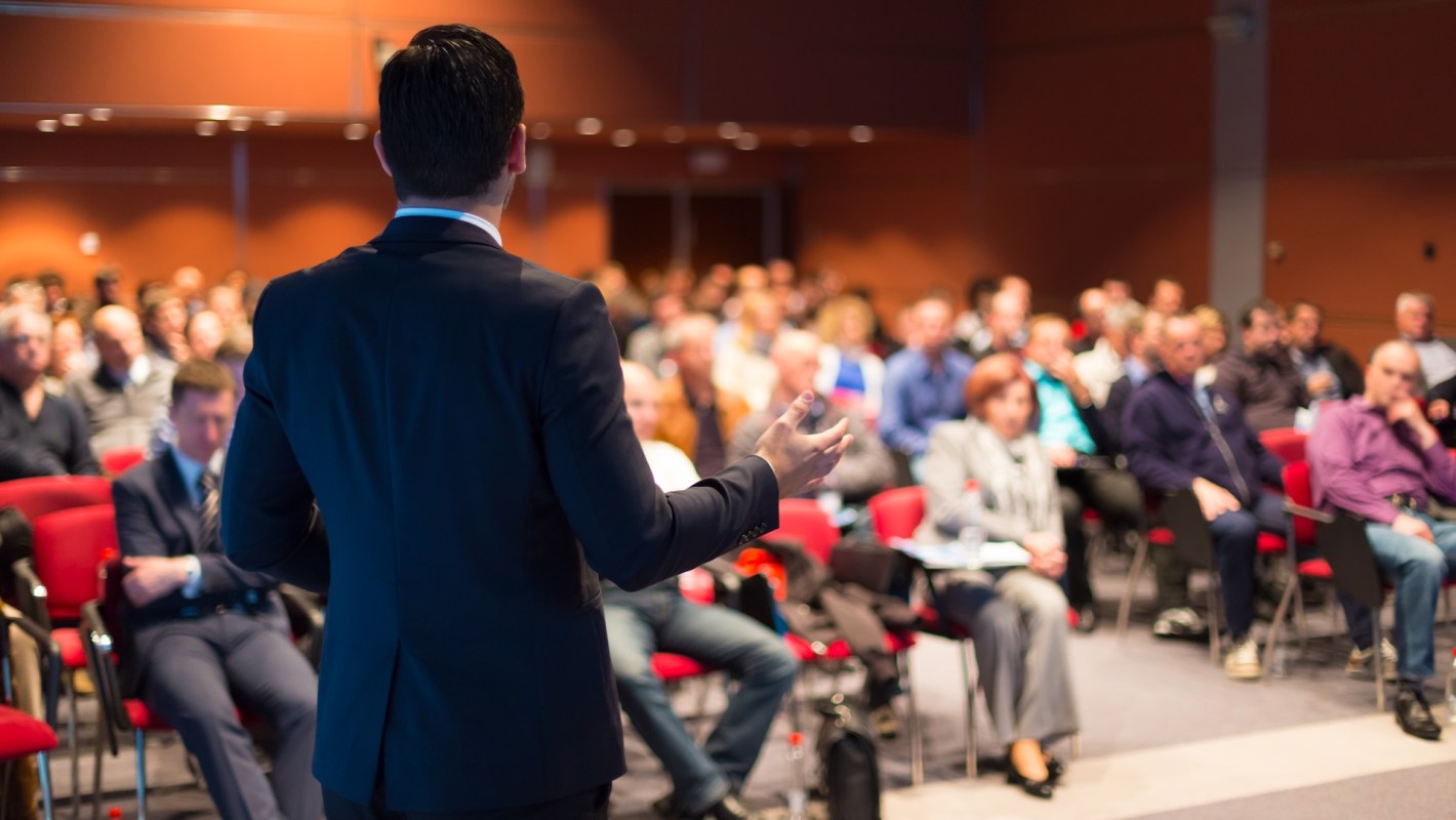 A speaker presents to an audience seated in a conference room.