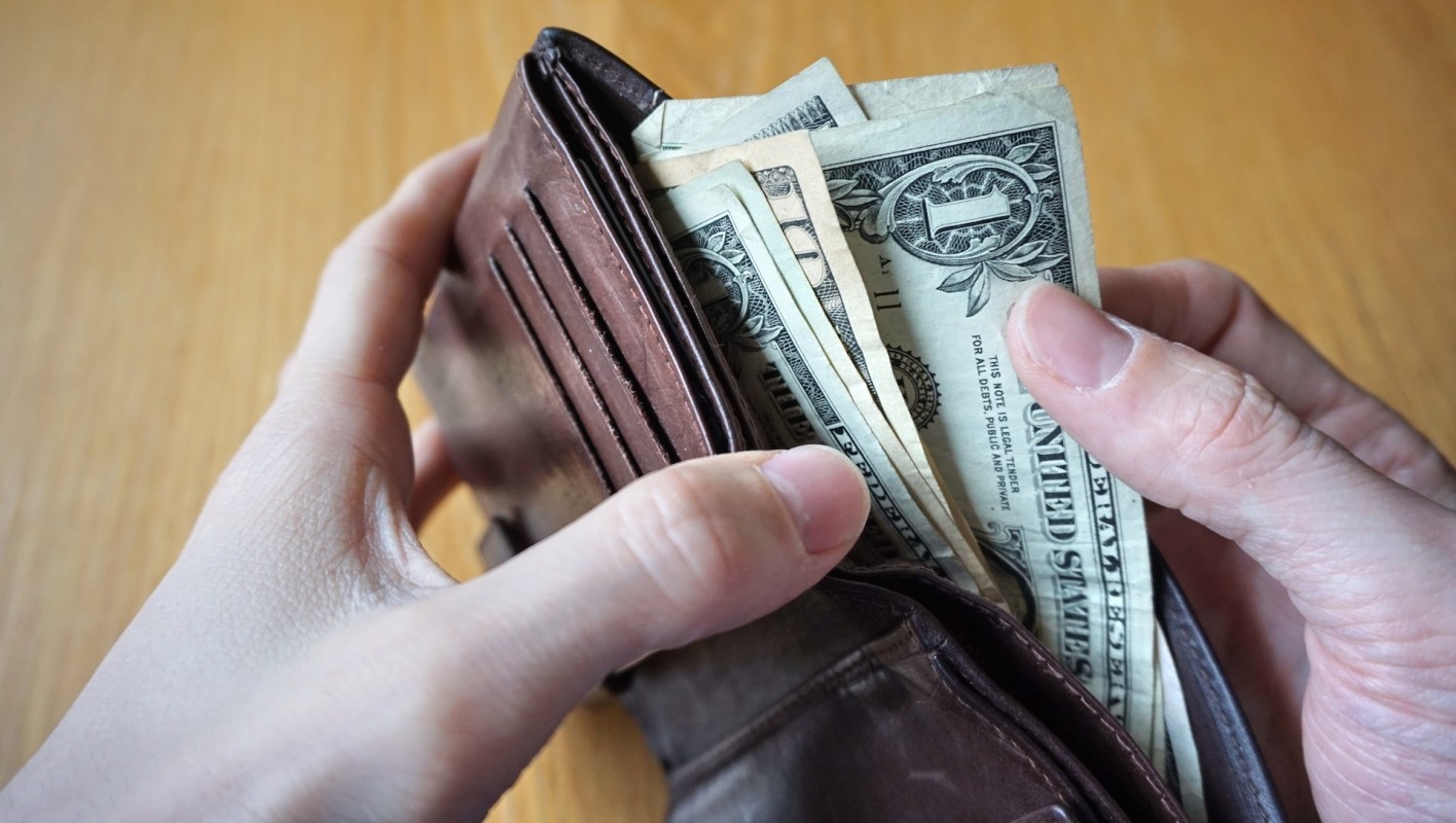 A person holding a brown wallet with US dollar bills, including $1 and $10 bills, inside.