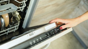 A hand is pressing a button on a dishwasher with dishes visible inside.