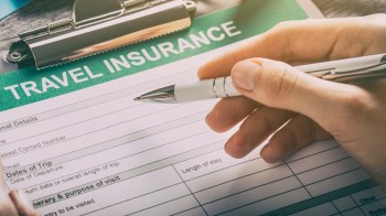 A hand holds a pen above a travel insurance form with various fields to fill out.