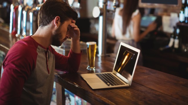 A man sits at a bar with a laptop and a beer, resting his forehead on his hand.