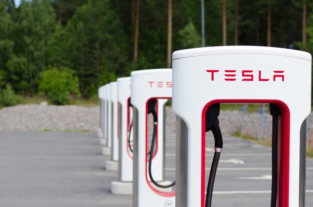 Multiple Tesla electric vehicle charging stations lined up in a parking area surrounded by trees.