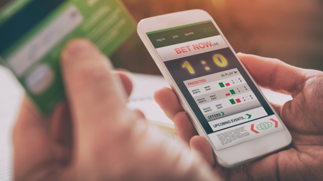 A person holds a smartphone showing a sports betting application with live scores and betting options.
