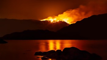 A mountain is illuminated by a fire against a dark sky, reflecting orange light on a calm body of water.