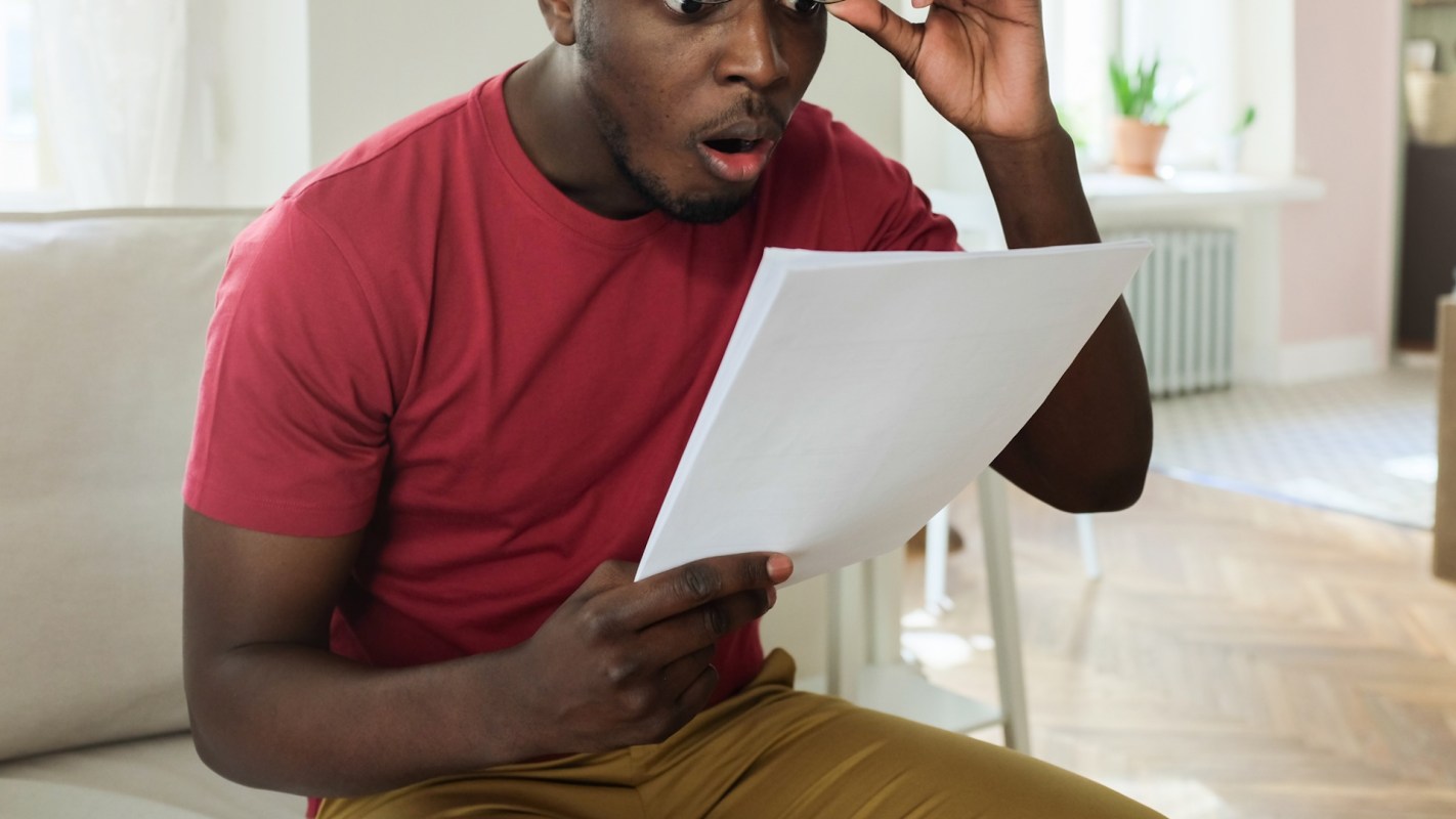 A man reading a piece of paper with his eyes wide open and expressing shock while sitting on a sofa.
