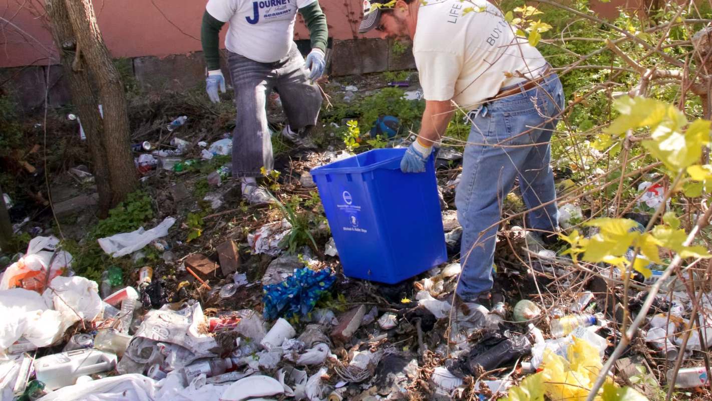 Two individuals clean up a site filled with trash and debris, using a blue recycling bin.