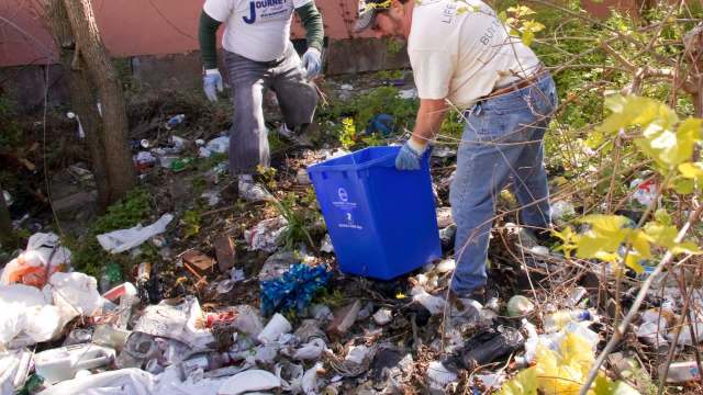 Two individuals clean up a site filled with trash and debris, using a blue recycling bin.