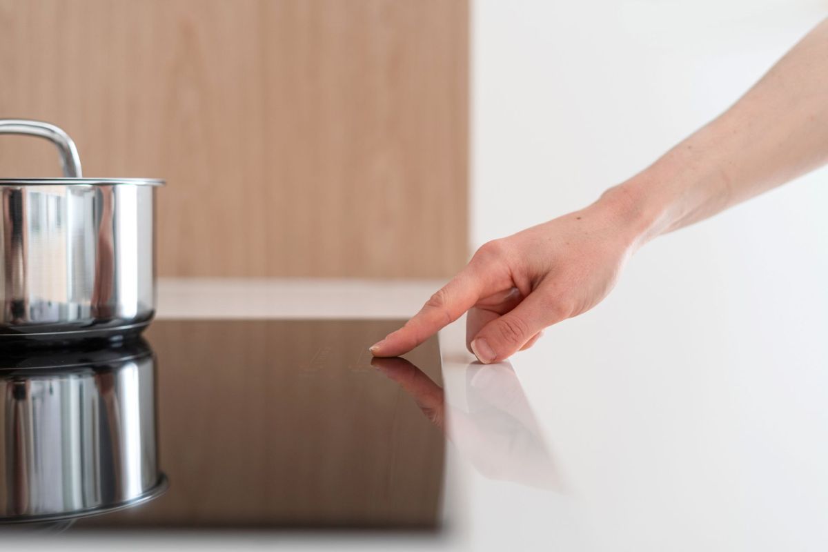 A hand touches a black induction cooktop next to a stainless steel pot.