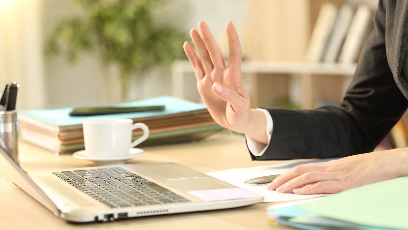 A hand raised in a greeting gesture beside a laptop and a cup on a table with various files in the background.