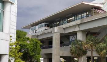 A monorail train is stationed at a elevated transit station surrounded by greenery and modern architecture.