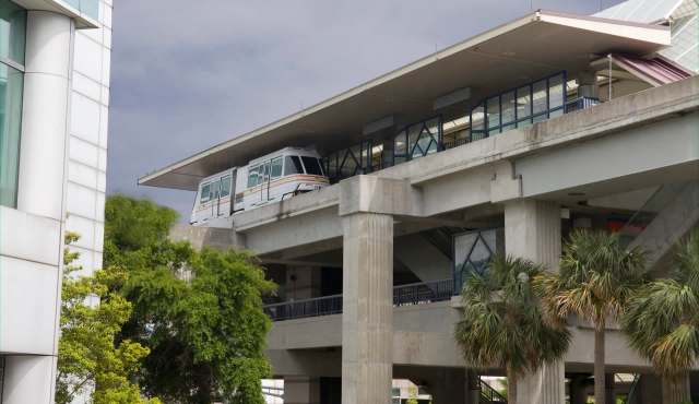 A monorail train is stationed at a elevated transit station surrounded by greenery and modern architecture.