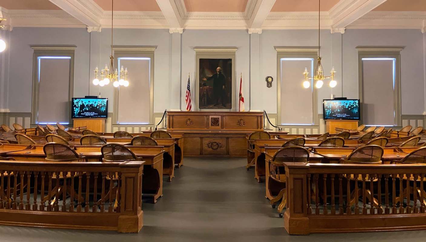 A historic courtroom with wooden furniture, American and Florida flags, and a television displaying a black-and-white image.