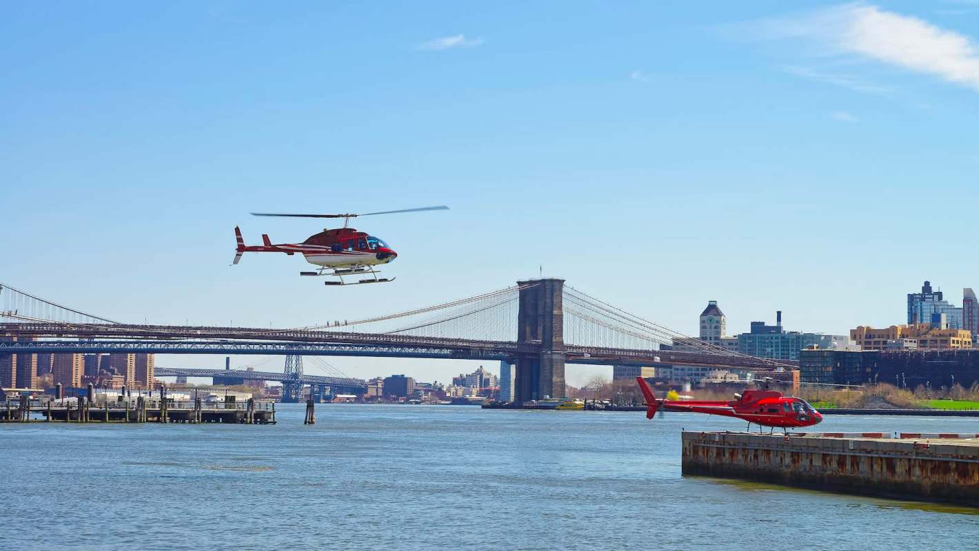 Two helicopters fly near the water with the New York City skyline and bridge in the background.