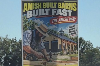 A promotional sign featuring an Amish carpenter building a barn with text announcing fast construction services.