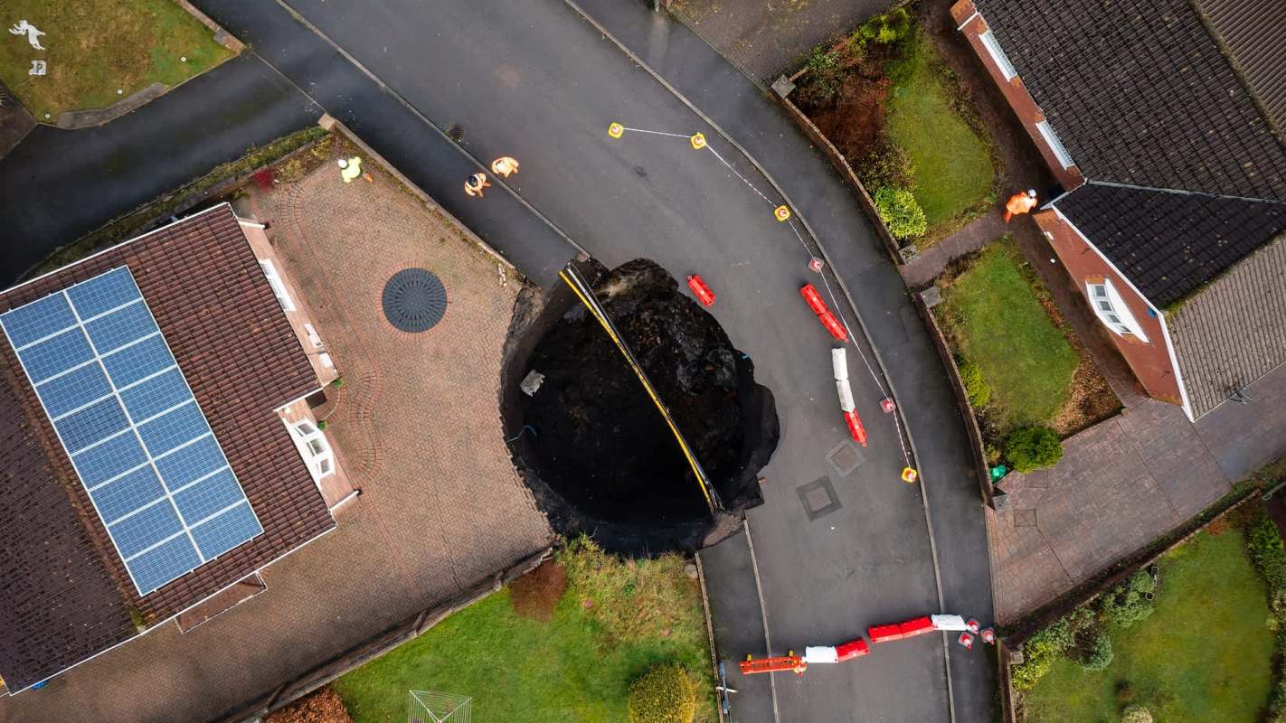 A large sinkhole has formed in a residential street, surrounded by traffic cones and construction barriers.