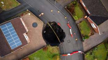 A large sinkhole has formed in a residential street, surrounded by traffic cones and construction barriers.