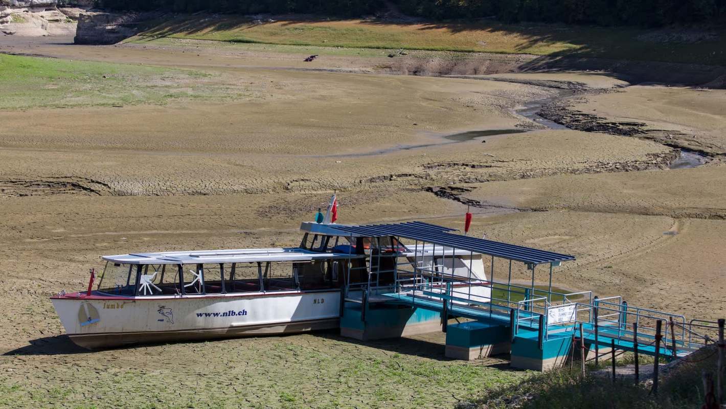 A boat docked on dry land at Les Brenets, Switzerland, with cracked earth and sparse vegetation surrounding it.