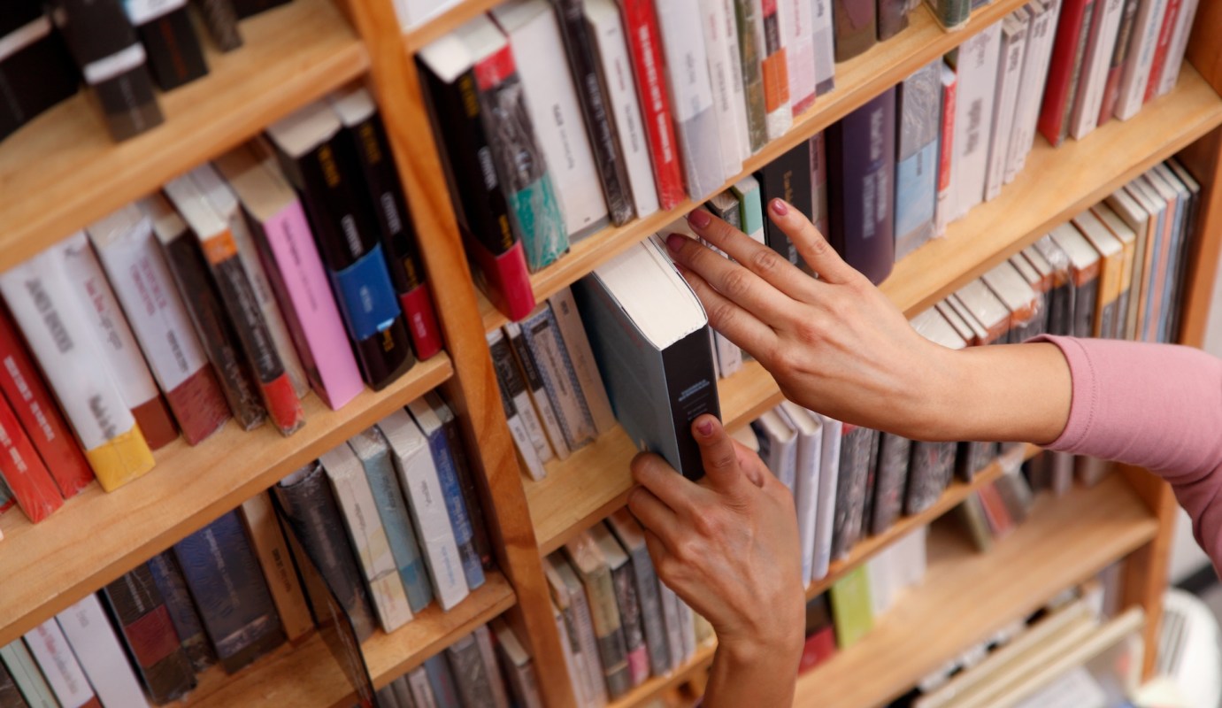 A person placing a book back on a wooden bookshelf filled with various books.