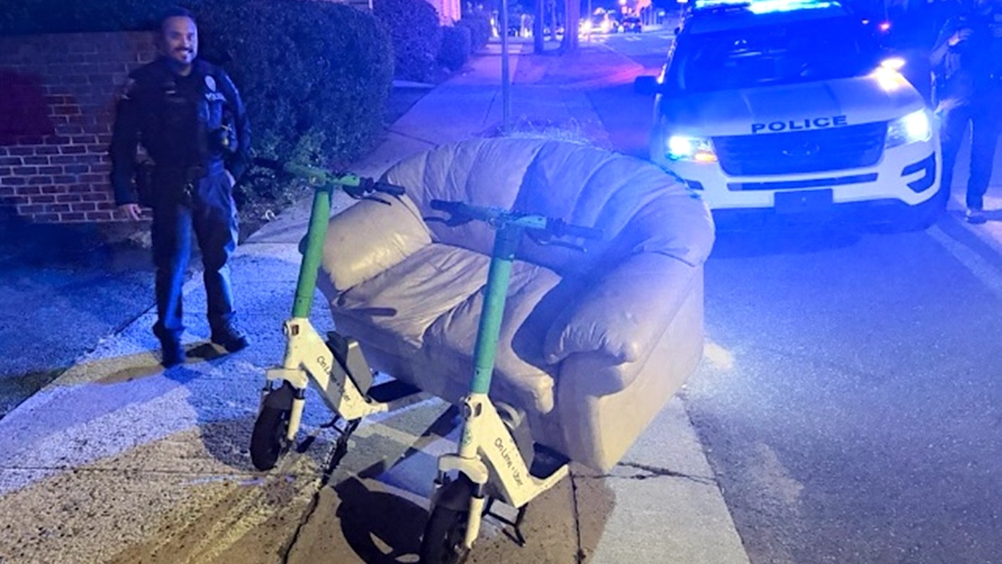 A police officer stands next to a couch attached to two electric scooters on a city street.