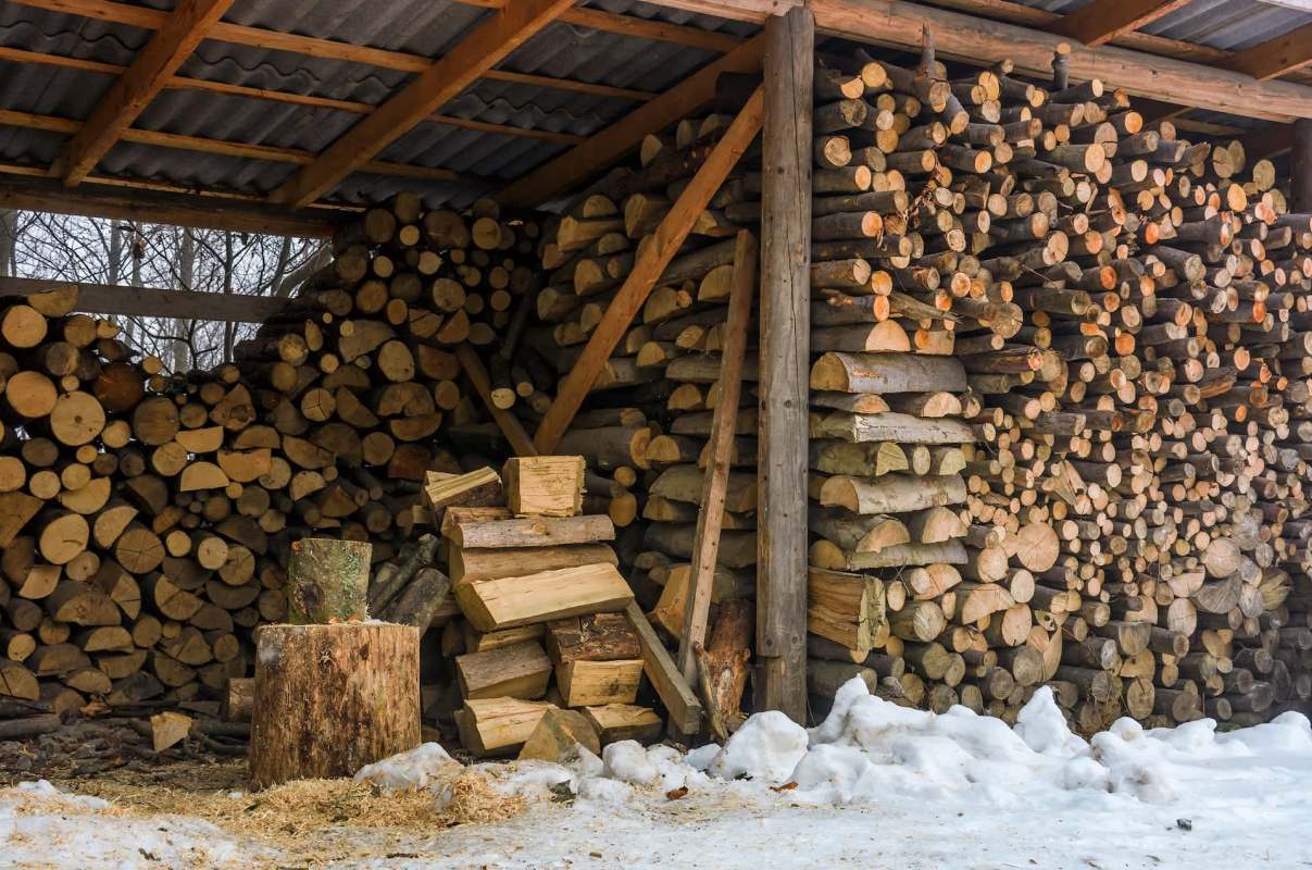 A woodpile storage area with stacked logs and pieces of timber on snow-covered ground.