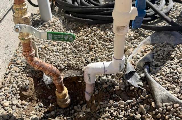 Pipes and fittings made of copper and PVC laid on gravel, with a partially dug hole in the ground.