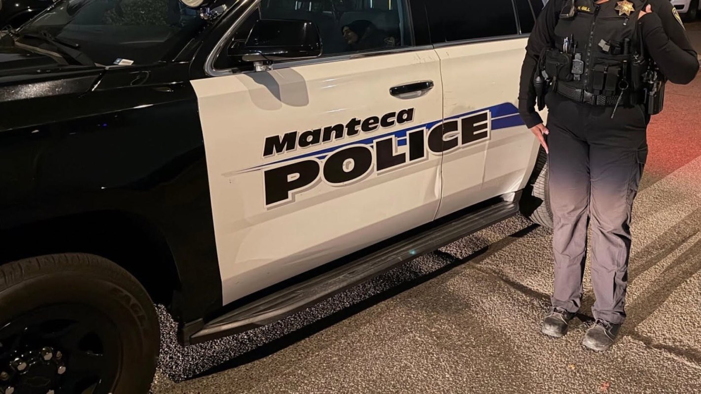 A police officer stands next to a marked police vehicle with "Manteca Police" displayed on the side.