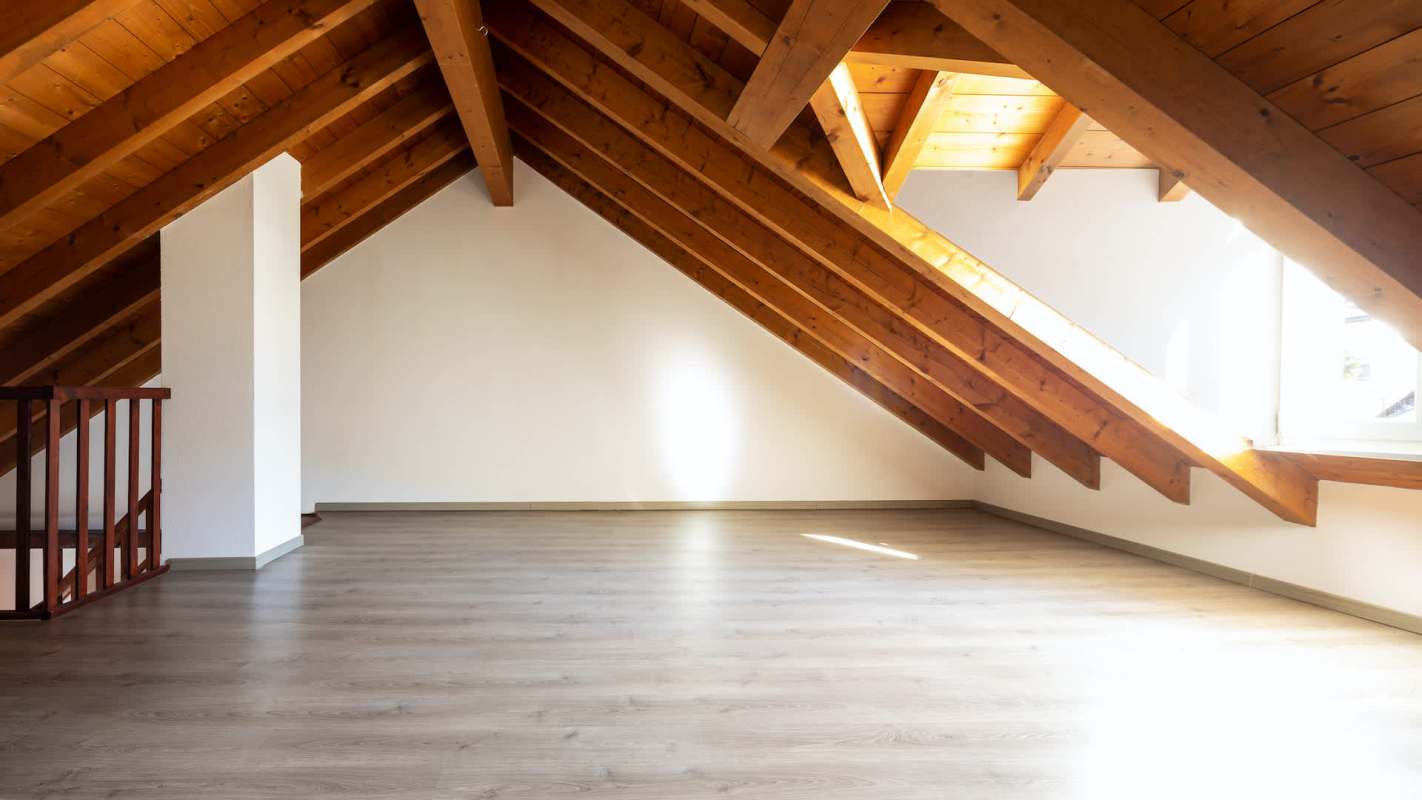 An empty attic space with wooden beams, a light-colored floor, and a window letting in natural light.