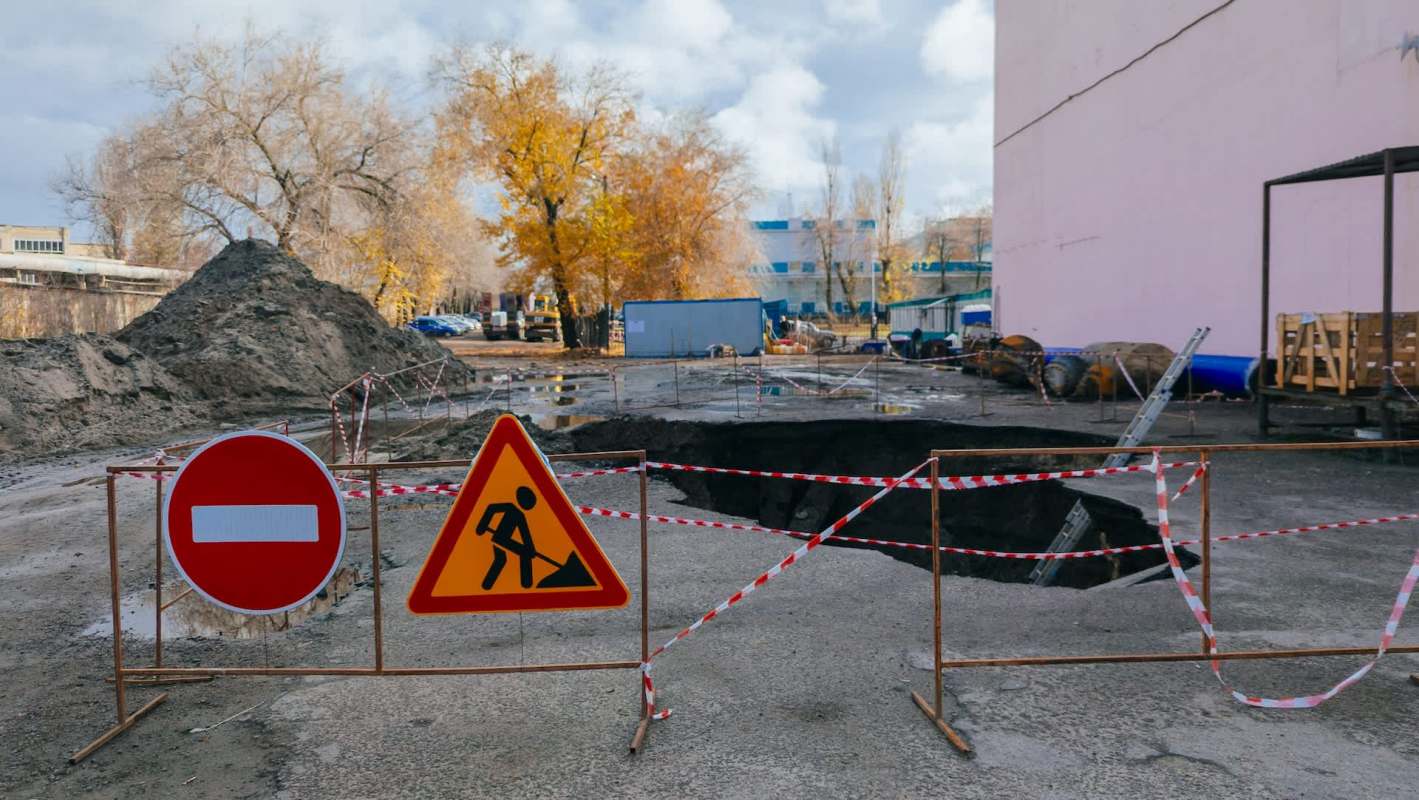 A construction site with a large hole in the ground, marked by warning signs and barriers.