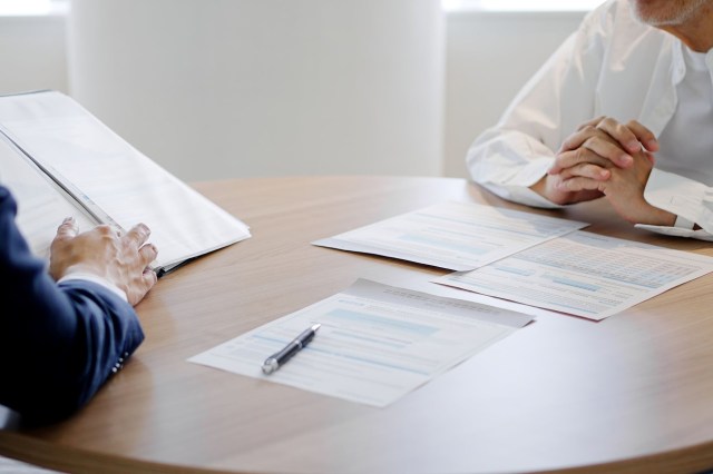 Two people sit at a table with documents and a pen between them, engaged in discussion.
