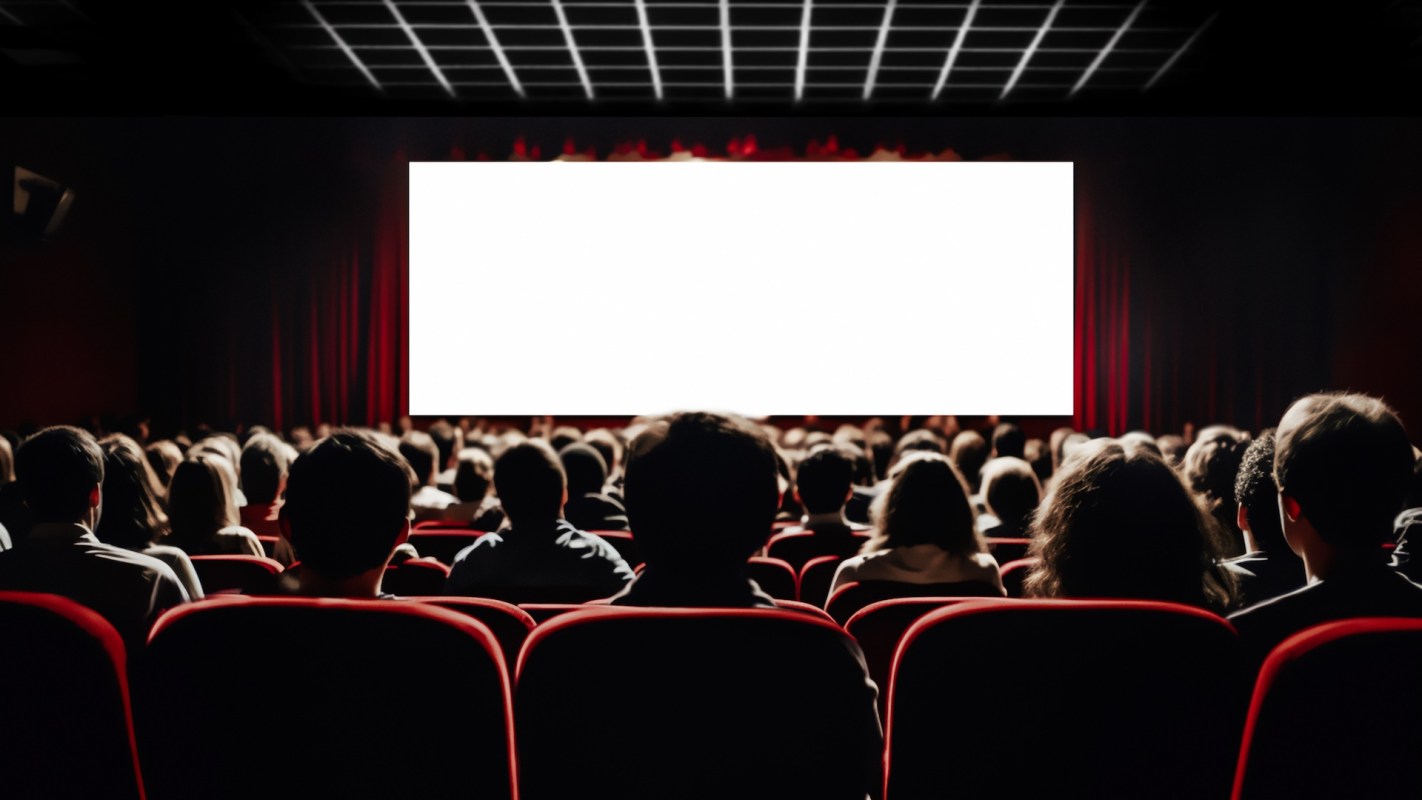An audience seated in a theater facing a blank screen with red curtains.